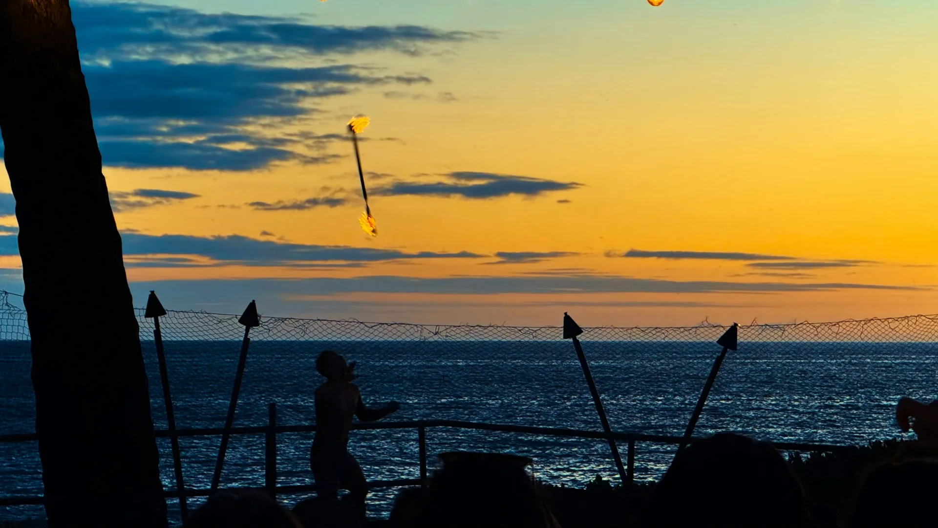 A fire-knife dancer silhouetted against an orange sunset over the ocean at Kailua Bay, tiki torches lit along the shoreline rail
